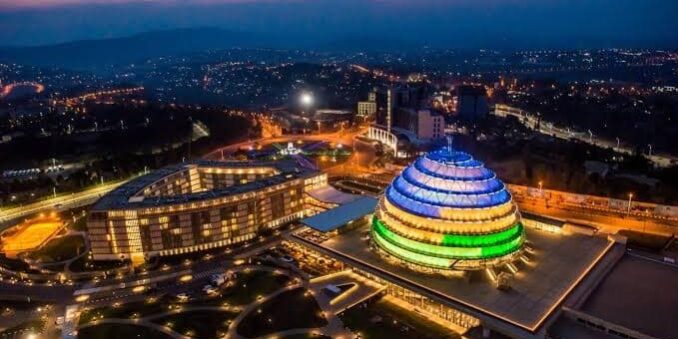 Aerial night view of the Kigali Convention Centre in Rwanda, illuminated in vibrant blue, yellow, and green lights, symbolizing the national flag colors.
