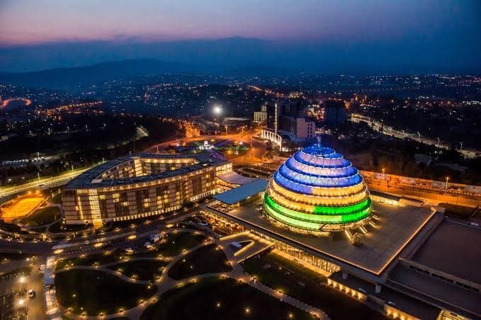 Aerial night view of the Kigali Convention Centre in Rwanda, illuminated in vibrant blue, yellow, and green lights, symbolizing the national flag colors.