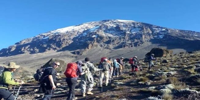 Trekkers ascending Mount Kilimanjaro with snow-capped peak in view under a clear blue sky – GoFurther4x4CarRental