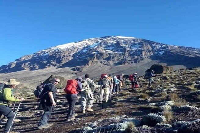 Trekkers ascending Mount Kilimanjaro with snow-capped peak in view under a clear blue sky – GoFurther4x4CarRental